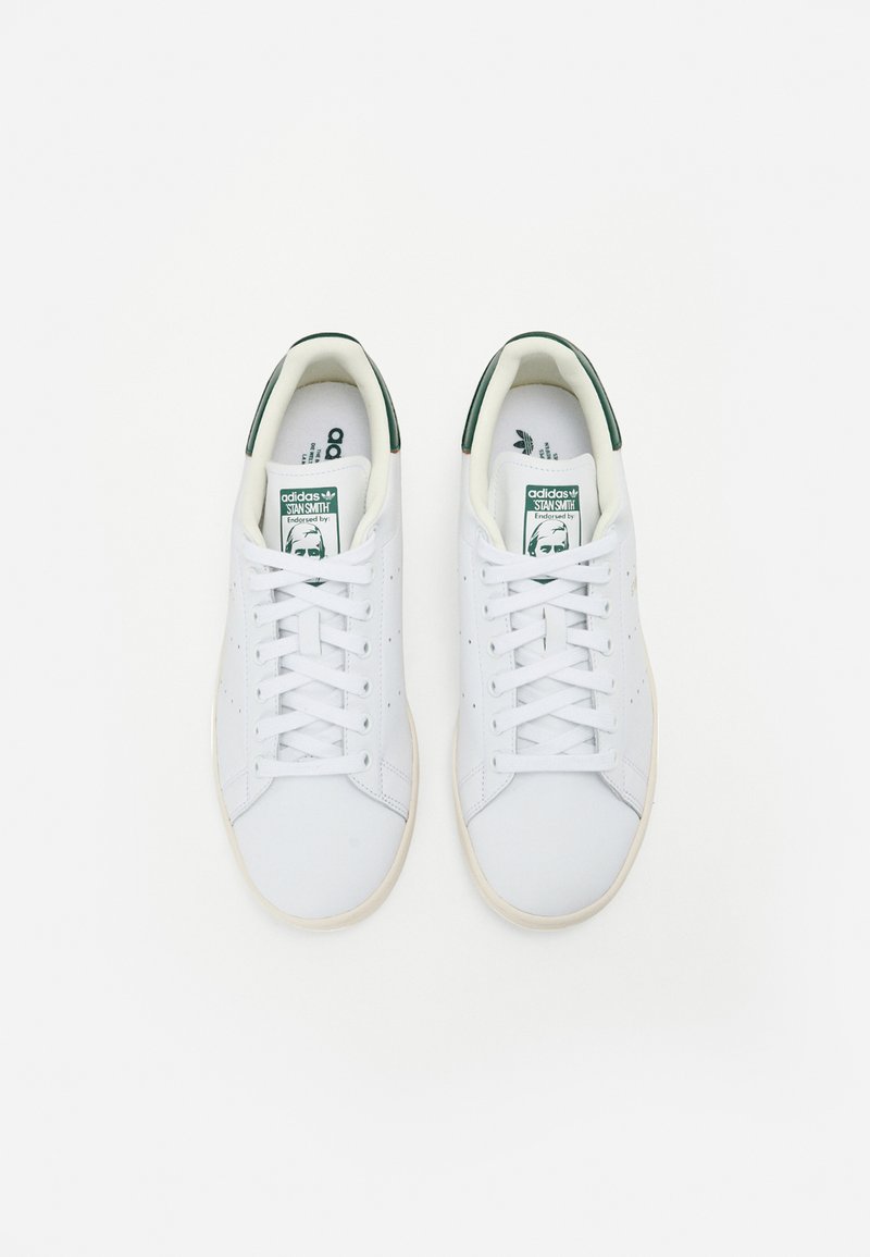 Baskets en cuir blanc avec une texture lisse, accents verts sur le talon et la languette, dotées de lacets plats et de détails perforés sur les côtés.