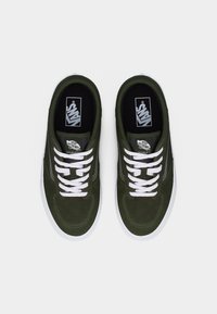 Par rjavo zelenih superg Vans z belimi vezalkami in belimi podplati, prikazanih od zgoraj na enostavni belem ozadju.