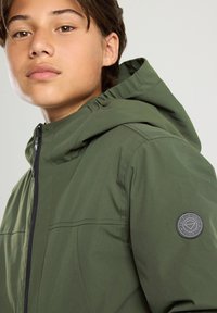 Teen wearing a green hooded jacket with a zipper and a round "America Today Iconic" patch on the sleeve, looking forward.