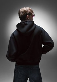 Černá mikina s kapucí a hladkou texturou, s volným střihem. Osoba má na sobě tmavé džíny a sluneční brýle, stojí zády k fotoaparátu.