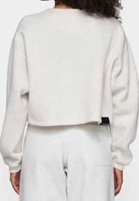 Persona che indossa un cropped sweatshirt beige chiaro e pantaloni della tuta abbinati, visto da dietro su uno sfondo bianco semplice.