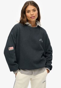 Felpa oversize blu navy con collo a coste, con logo frontale e patch della bandiera americana sulla manica sinistra. Maglietta a righe sotto.