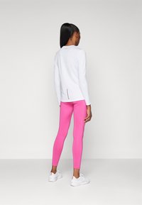 Bela majica s dolgimi rokavi in razporkom na hrbtni strani, v kombinaciji s svetlo roza leginami. Manekenka nosi bele čevlje z oranžnimi poudarki. Gladka tekstura tkanine.