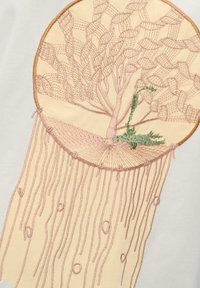 Geborduurde kunst met een boom van roze en groene draden op een crème kleurige achtergrond, omringd door decoratieve franjes.