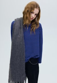 Jonge vrouw met golvend blond haar, draagt een blauw gestreept shirt en zwarte broek, met een lange grijze sjaal over één schouder gedrapeerd.