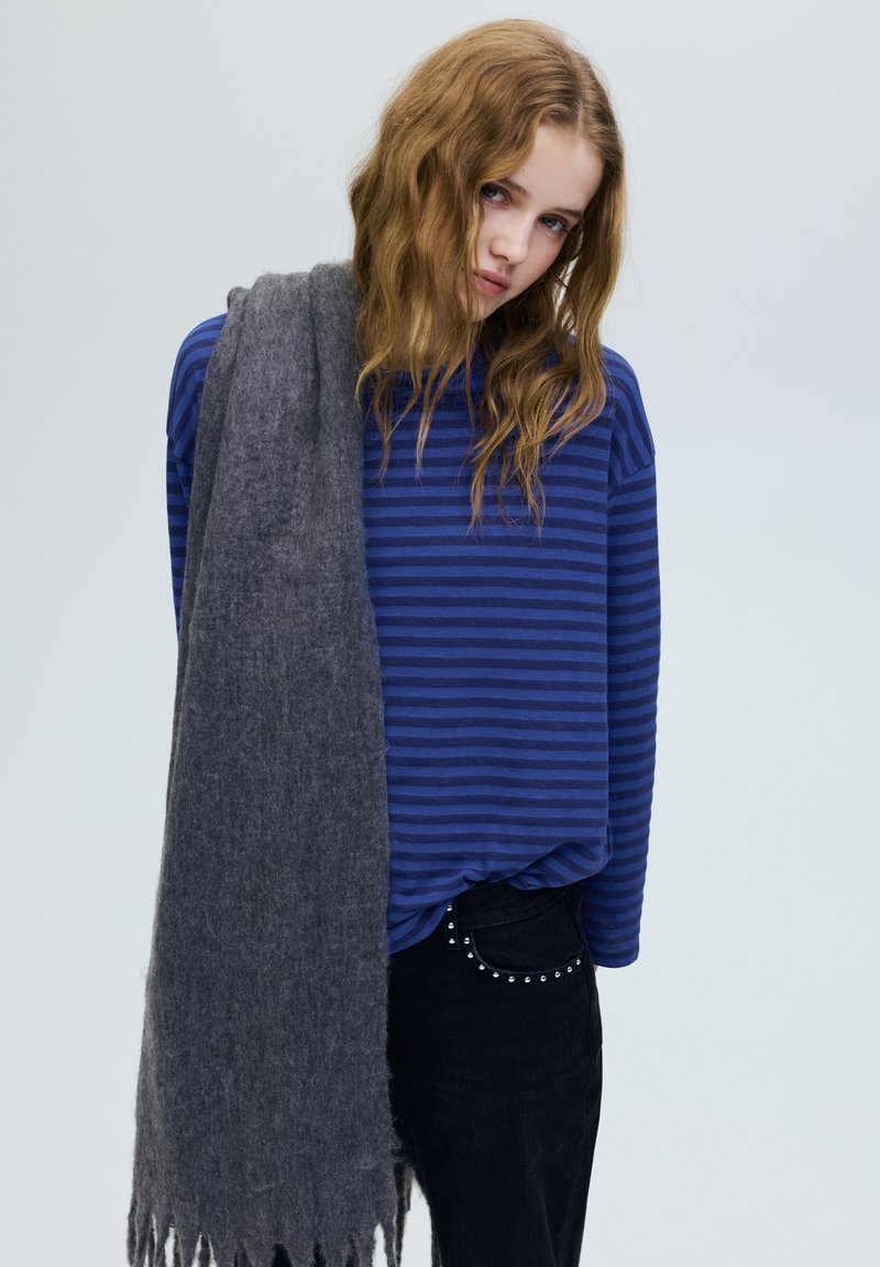 Jonge vrouw met golvend blond haar, draagt een blauw gestreept shirt en zwarte broek, met een lange grijze sjaal over één schouder gedrapeerd.