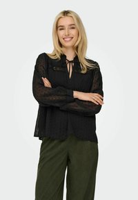 Schwarze durchsichtige Bluse mit Punkten, langen Ärmeln, einem Bindeband am Hals und Spitzenverzierungen. Kombiniert mit grünen Cordhosen.