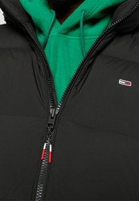 Svart pufferväst med dragkedja över en grön hoodie. Har en logotypapplikation och en dragkedja med röda och vita detaljer. Strukturerat tyg.