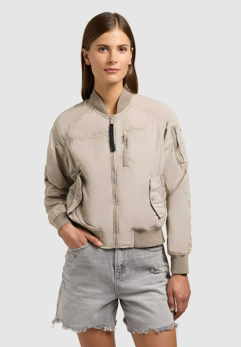 Žena v béžové bomber bundě se zipem a kapsami, která má na sobě světle šedé roztrhané riflové šortky, stojící na neutrálním pozadí.