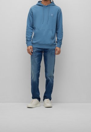 Mężczyzna w niebieskiej bluzie z kapturem, średnio pranych niebieskich dżinsach i białych trampkach stoi na tle jednolitego jasnoszarego tła.