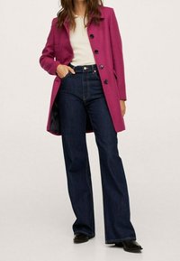 Abrigo fucsia con cuello de muesca y botones negros, combinado con jeans oscuros de tiro alto y pernera acampanada, y botas negras hasta el tobillo.