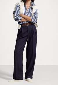 Mörkblå vida byxor i kombination med en blå och vit randig skjorta och en krämfärgad stickad väst. Brun bälte med ett guldspänne, vita sneakers.