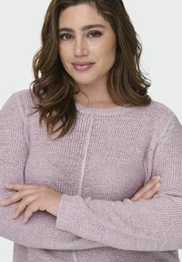 Heled roosa kudumiskoht, millel on tekstuurne kudumine, ümmargune kaelus ja soonikkoes varrukad. Mudel on käed ristis ja näeb välja rahulik.