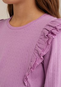 Purple ribbed sweater featuring a round neckline and a ruffled accent on the left shoulder, made from textured knit fabric.