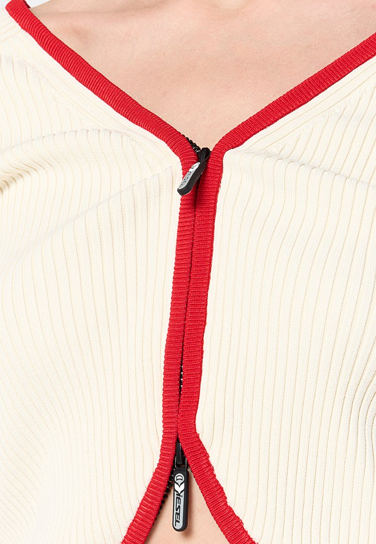 Top a costine color crema con scollatura a V profonda, caratterizzato da una zip rossa prominente e rifiniture rosse lungo i bordi. Tessuto leggero.