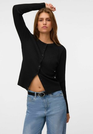 Jeune femme en cardigan noir boutonné et jean bleu pose avec un bras levé sur un fond blanc uni.