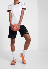 Persona vestida con camiseta deportiva blanca, pantalones cortos negros, calcetines blancos y zapatillas de deporte naranja-rosa, sosteniendo una pelota de balonmano naranja y rosa.