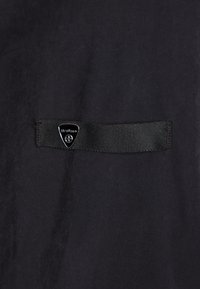 Czarny tekstylny materiał o gładkiej fakturze z prostokątnym czarnym paskiem i trójkątną metką z logo z białym napisem i emblematem.