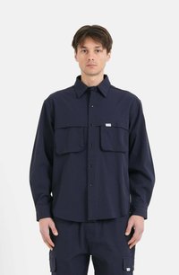 Camicia a maniche lunghe blu navy con colletto, due tasche frontali e bottoni a pressione, realizzata in un tessuto uniforme e liscio. Senza motivi visibili.