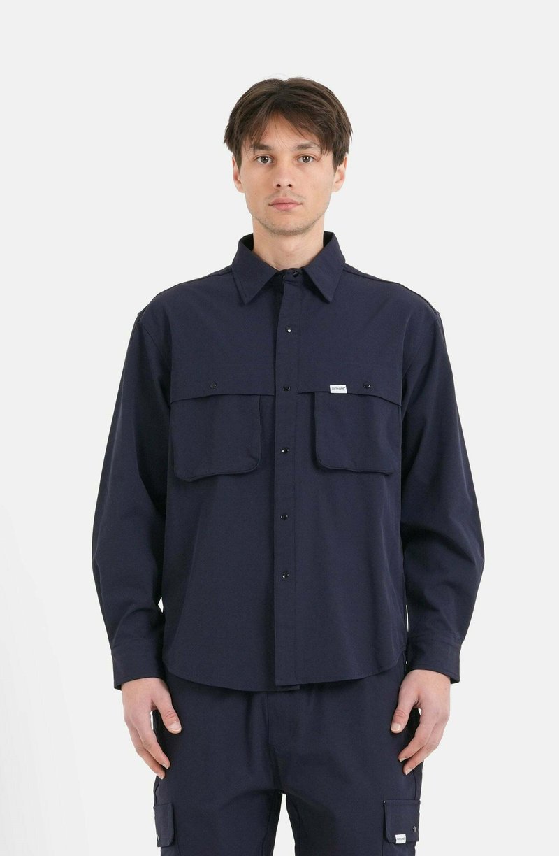 Camicia a maniche lunghe blu navy con colletto, due tasche frontali e bottoni a pressione, realizzata in un tessuto uniforme e liscio. Senza motivi visibili.