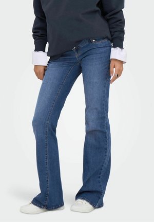 Osoba nosząca niebieskie jeansy typu bootcut, białe trampki i ciemny długi rękaw z białymi mankietami, stojąca na jednolitym tle.