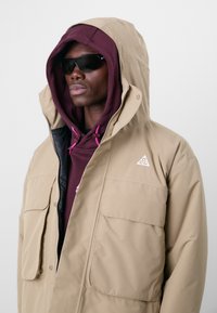 Beige puffer jacket with a hood, featuring large pockets and a logo on the chest. Underneath, a maroon hoodie with pink accents. Black sunglasses.