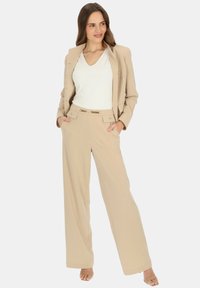 Beżowy garnitur na miarę z dopasowaną marynarką i szerokimi spodniami. Biała koszula z dekoltem w serek pod spodem. Metalowy akcent na pasku, gładka faktura materiału.
