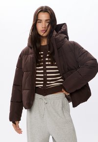 Brązowa kurtka puchowa z kapturem, z pikowanym wzorem i elastycznymi mankietami, noszona na paskowanym, zapinanym na guziki topie oraz szarych dresach.