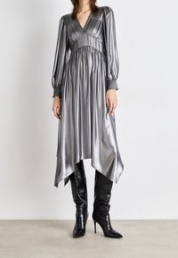 Robe argentée métallique courte devant, longue derrière, avec des manches bouffantes et un buste plissé. Associée à des bottes noires à talons aiguilles montantes jusqu'aux genoux. Texture lisse.