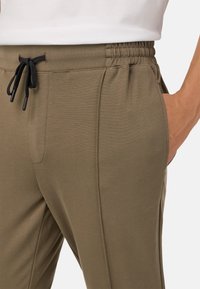 Žalios alyvuogių spalvos elastingi kelnės su juodu raišteliu, šoninėmis kišenėmis ir lygia tekstūra, turinčios siaurėjantį dizainą.