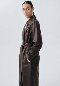 Długi brązowy skórzany płaszcz z paskiem w talii, bocznymi kieszeniami i klapami, charakteryzujący się gładką teksturą i lekko otwartym przodem.