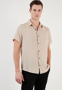 Korte mouw beige shirt met een kraag, textuurstof en donkerblauwe knopen, gedragen met zwarte broek.