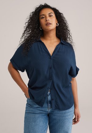 Kvinde med krøllet hår iført en løs navyblå tekstureret skjorte med knapper og blå jeans, stående med den ene hånd i lommen.