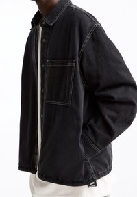 Sort denimjakke med knaplukning foran, brystlomme, synlige syninger og løs pasform. Stylet over en hvid skjorte.