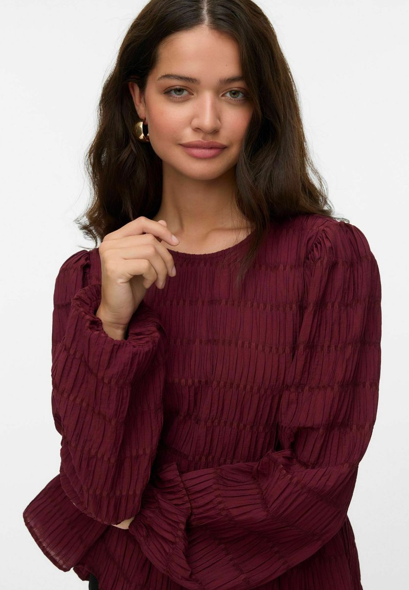 Haut texturé bourgogne avec un design plissé, encolure ronde et manches bouffantes, associé à des boucles d'oreilles rondes en or.
