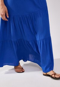 Abito lungo di un blu vivace, con un design a strati e tessuto testurizzato. Abbinato a sandali flat marroni.