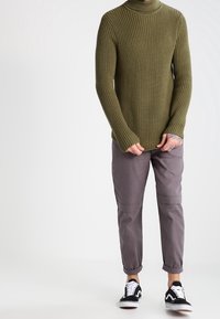 Maglione a collo alto a coste verde oliva con maniche lunghe, abbinato a pantaloni grigi a gamba stretta e sneakers nere con dettagli bianchi.