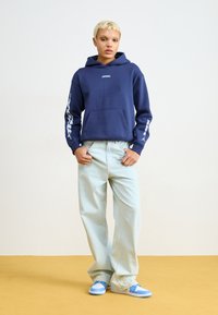 Sweatshirt azul-marinho com bolso frontal e logo branco nas mangas, combinado com jeans de perna larga azul claros e sapatilhas azuis sobre um chão amarelo.