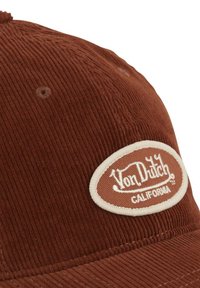 Casquette en velours côtelé marron avec un patch ovale blanc affichant le texte "Von Dutch California". Elle possède une visière structurée et une surface texturée.