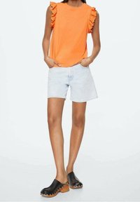 Haut orange sans manches avec des accents à volants, associé à un short en denim bleu clair et des sabots noirs avec talons en bois et clous métalliques.