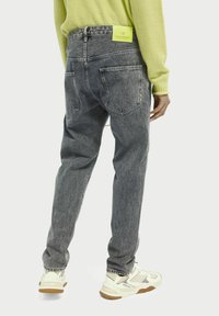 Szare jeansy z denimu o luźnym kroju, z pięcioma kieszeniami, widocznymi przeszyciami i żółtą, markową metką na pasku. Buty są białe z akcentami kolorystycznymi.