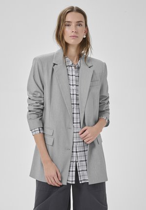 Femme aux cheveux mi-longs portant un blazer gris clair surdimensionné par-dessus une chemise à carreaux blanche et grise et un pantalon foncé, sur un fond uni.