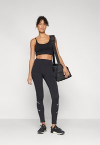 Soutien-gorge de sport noir, leggings noirs taille haute avec des accents blancs, et baskets noires. Le mannequin porte un sac noir et se tient devant un fond neutre.