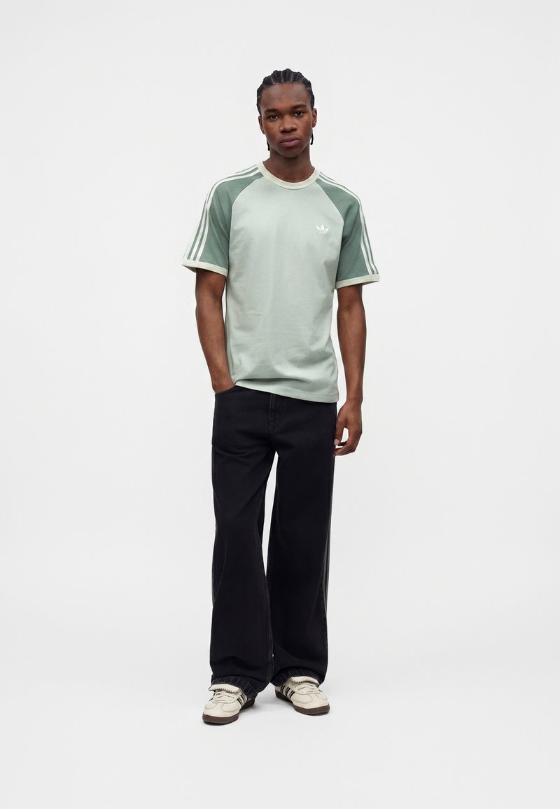 Ung mand stående, iført lysegrøn og hvid Adidas kortærmet skjorte, sorte bukser med vide ben og hvide sneakers med sorte striber.