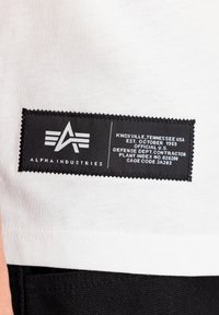 Witte stof met een zwart etiket met gestikte randen, waarop het logo "A" en tekst over het merk en zijn oorsprong staat.