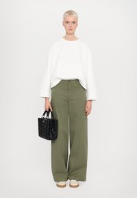 White long-sleeve sweatshirt with embroidered logo, olive wide-leg trousers, and a black woven handbag. Beige shoes complete the look.
