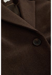 Brun ullblandningstyg av en kappa med en slitskrage och rund knappdetalj. Slät textur och subtil lyster i materialet.