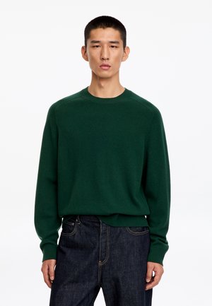 Homme portant un pull vert foncé à col rond et un jean bleu foncé, debout devant un fond blanc uni.
