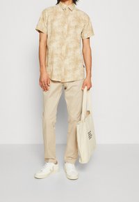 Chemise beige à manches courtes avec un motif de feuilles, associée à un pantalon beige clair et des baskets blanches. Porte un sac fourre-tout beige.