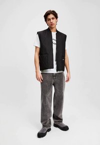 Zwarte gewatteerde vest met rits, met twee voorzakken, gedragen over een witte T-shirt met tekst. Grijze oversized jeans en zwarte schoenen.